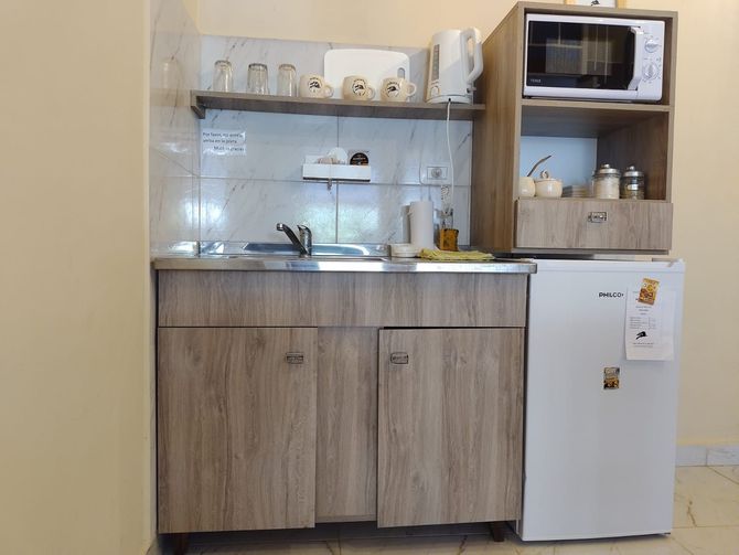 Pequeña cocina con gabinetes de madera clara, fregadero de acero inoxidable, microondas, mini refrigerador y estante con tazas.