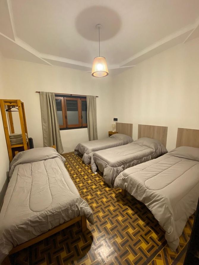 Cuatro camas en una habitación; ropa de cama gris claro, pisos de madera, ventana con cortinas, luz de techo.