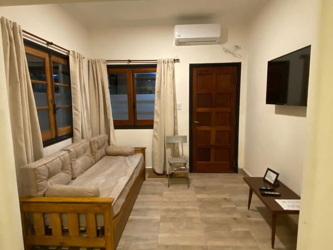 Acogedora sala de estar con sofá beige, puerta y molduras de madera, TV, ventana con cortinas y aire acondicionado.