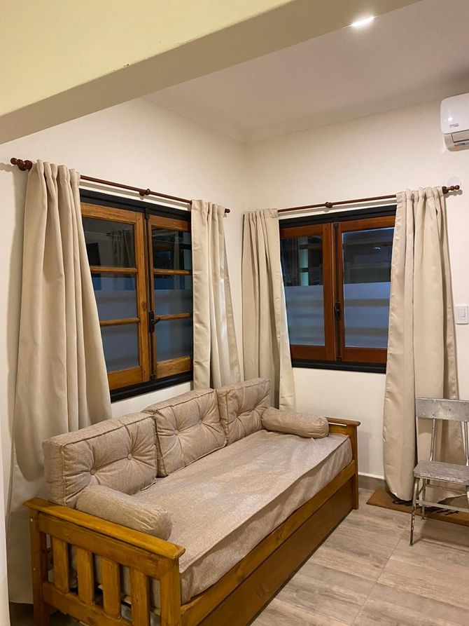 Rincón de estar: sofá de madera clara, dos ventanas con cortinas beige, paredes y suelo en tonos claros.