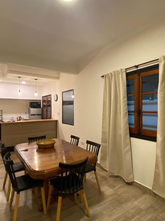 Comedor con mesa de madera y sillas negras, junto a una ventana con cortinas.