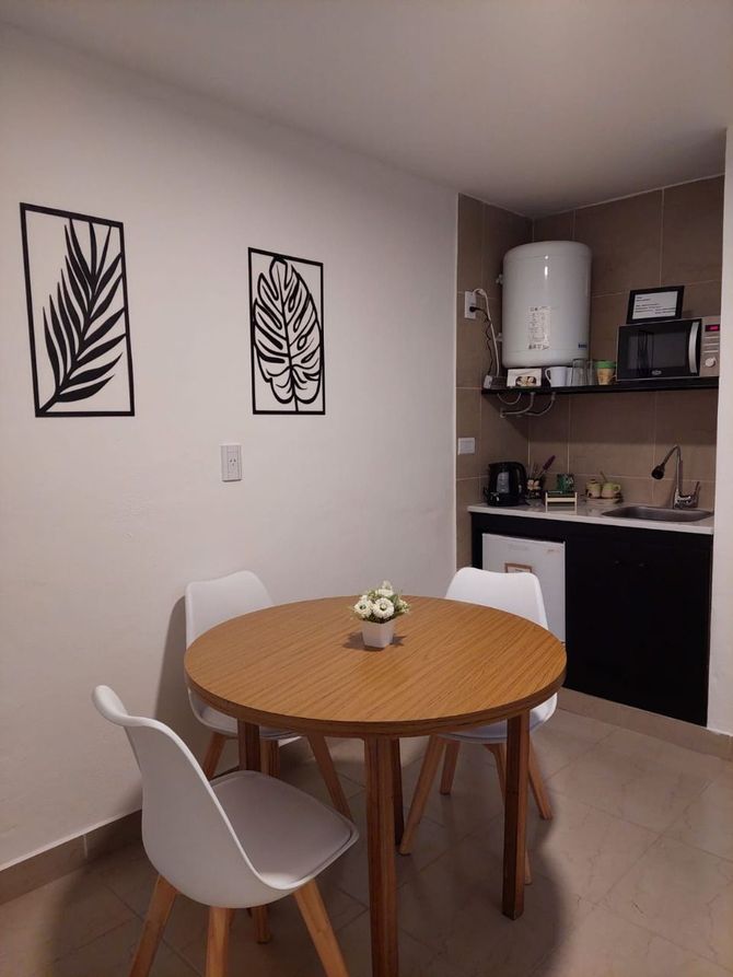 Un pequeño comedor con una mesa redonda de madera, tres sillas blancas y una pequeña cocina.