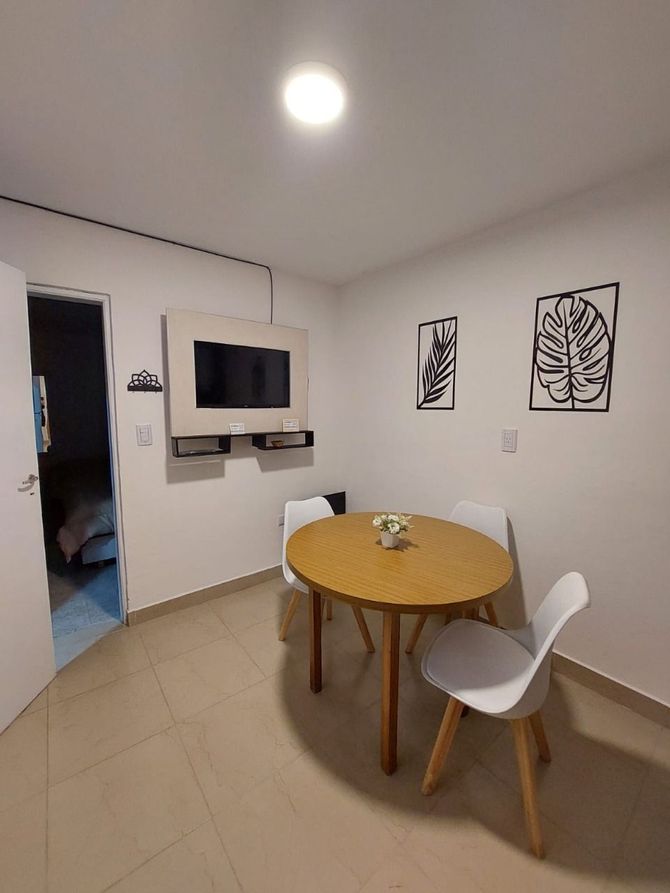 Pequeño comedor con mesa redonda de madera, sillas blancas y TV montada en la pared.