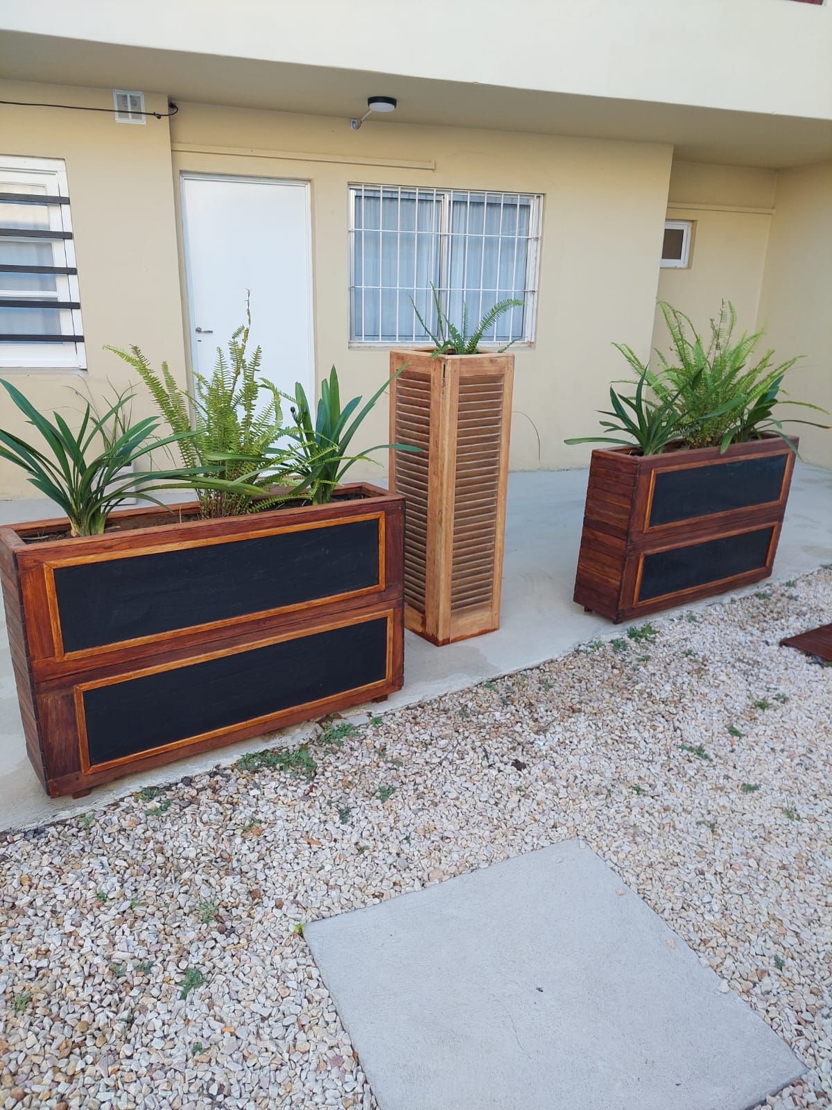 Tres jardineras de madera con vegetación en un patio de guijarros. Edificio beige al fondo.