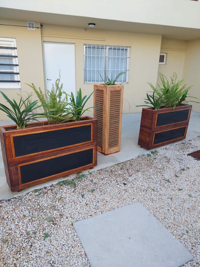 Tres jardineras de madera de distintos tamaños llenas de plantas en un patio de grava frente a un edificio.