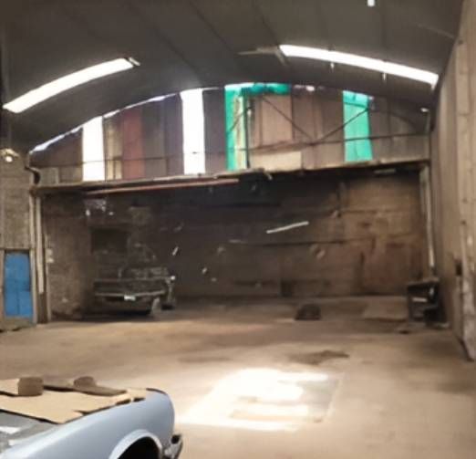 Interior de un gran almacén abierto y deteriorado con un automóvil, un techo expuesto y una puerta azul.