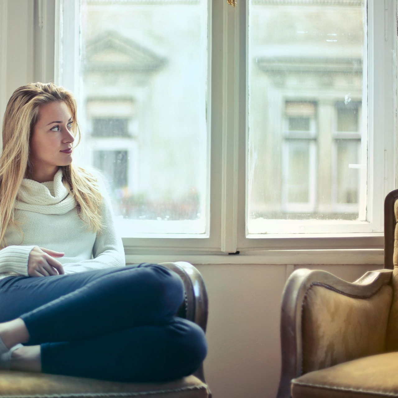Una mujer rubia con un suéter blanco está sentada junto a una ventana, mirando hacia afuera con una expresión pensativa.
