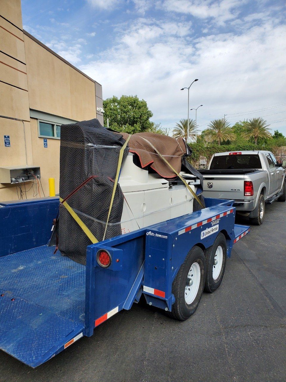 A pickup truck towing a blue trailer with a covered object secured by straps, parked near a building.