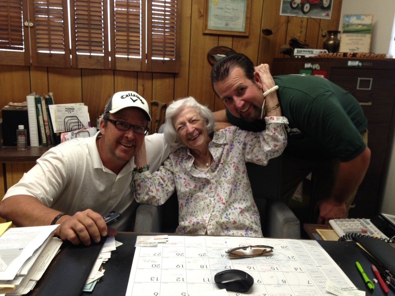 Two men flank an older woman smiling at a desk in an office.