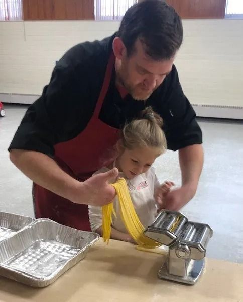 A man is teaching a little girl how to use a pasta machine