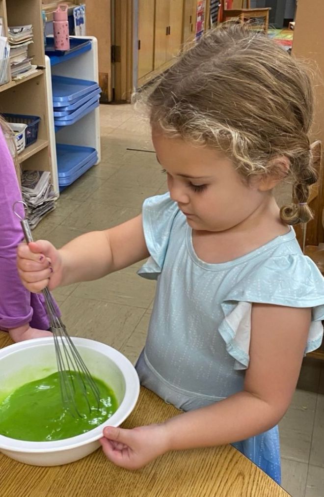 A little girl is mixing green liquid in a bowl with a whisk.