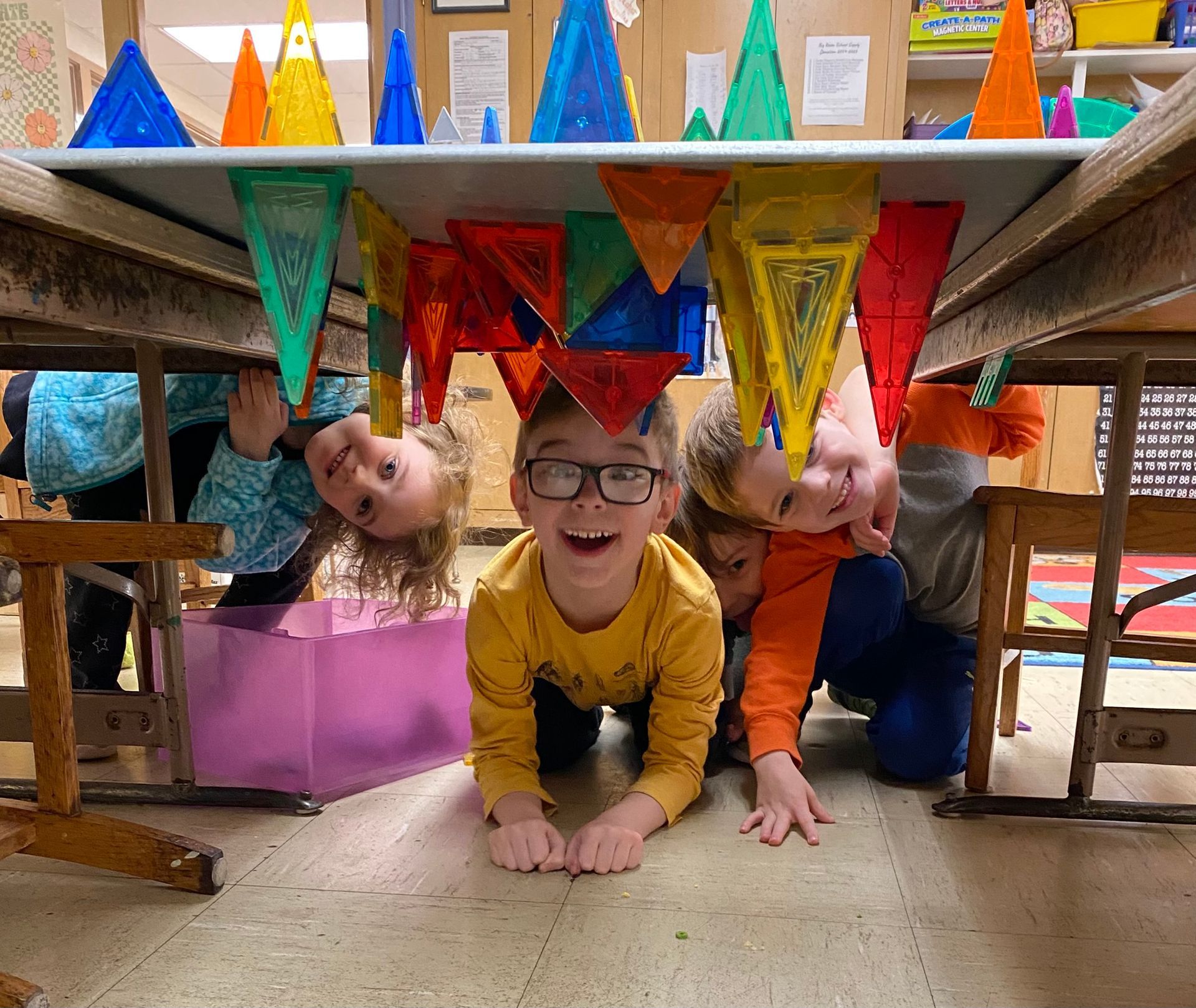 A group of children are playing under a table in a classroom.