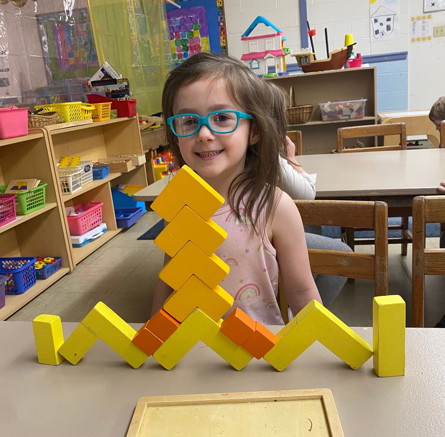 A little girl wearing glasses is sitting at a table with yellow and orange blocks