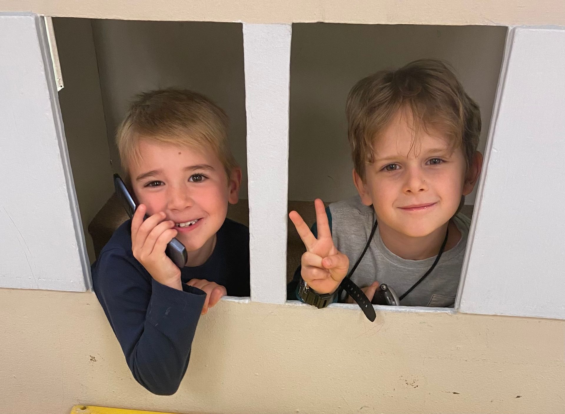 Two young boys are looking out of a cardboard box one is talking on a cell phone and the other is giving a peace sign