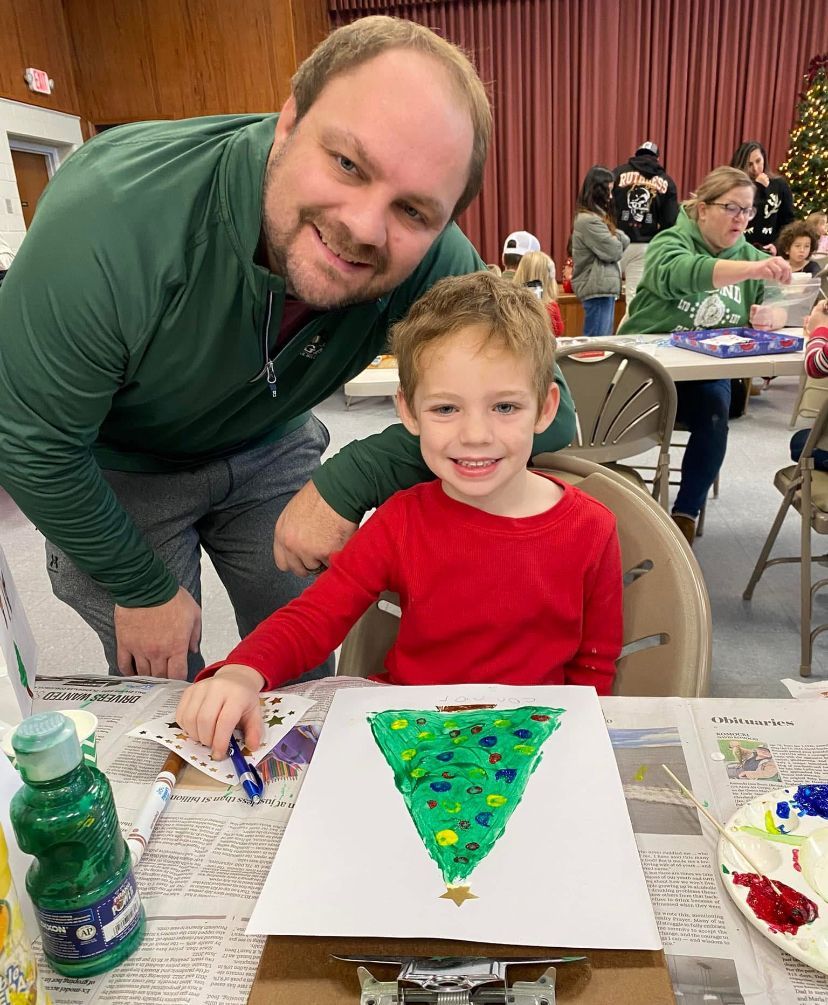 A man and a child are sitting at a table with a painting of a christmas tree.