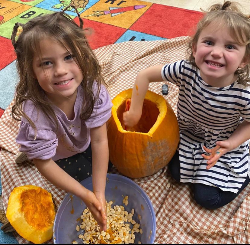 Two little girls are sitting on a blanket next to a pumpkin
