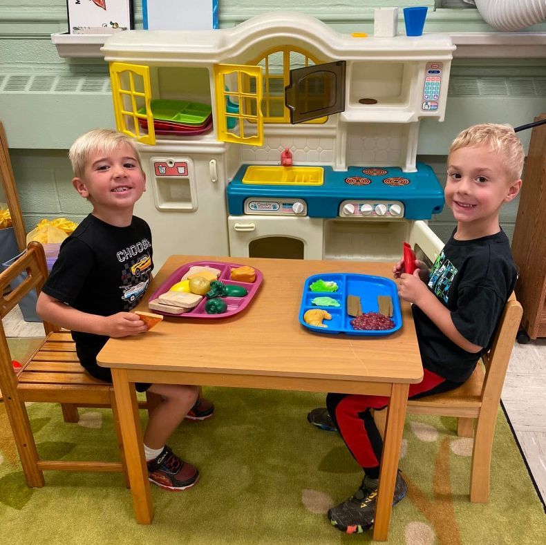 Two young boys are sitting at a table in front of a toy kitchen