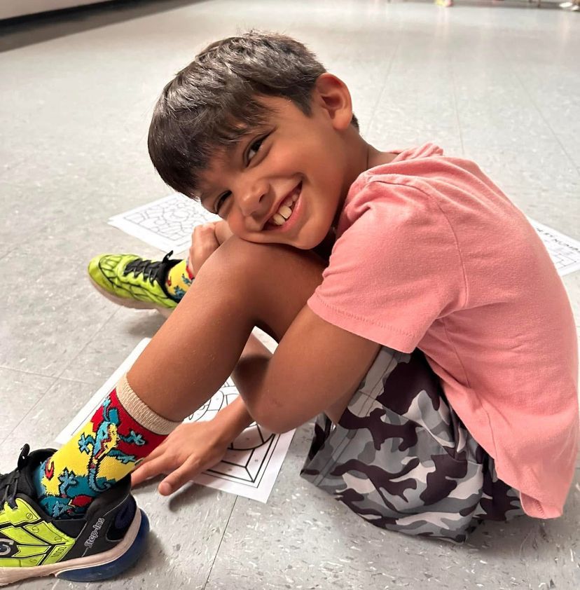 A young boy is sitting on the floor wearing a pink shirt and camouflage shorts
