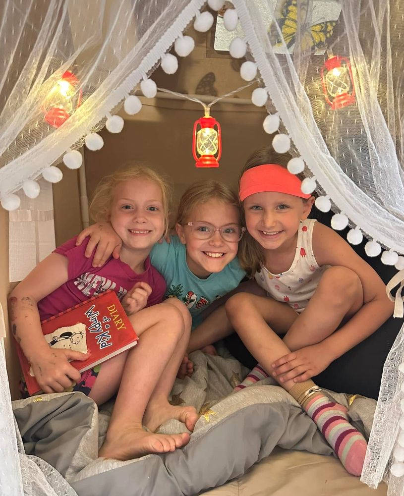 Three young girls are sitting under a canopy reading a book.