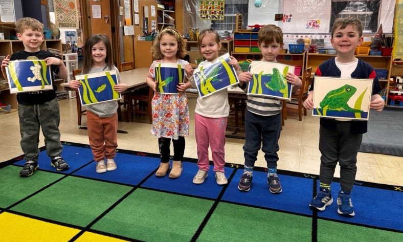 A group of children are standing next to each other in a classroom holding pictures of frogs.