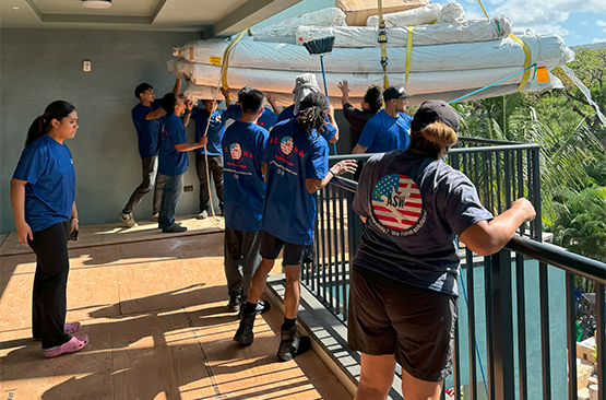 A group of people, many wearing blue shirts, lifting bundled items with ropes and pulleys onto a balcony overlooking trees.
