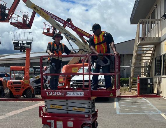 Two workers in safety vests stand in a red scissor lift. They are near a building with a parking area.