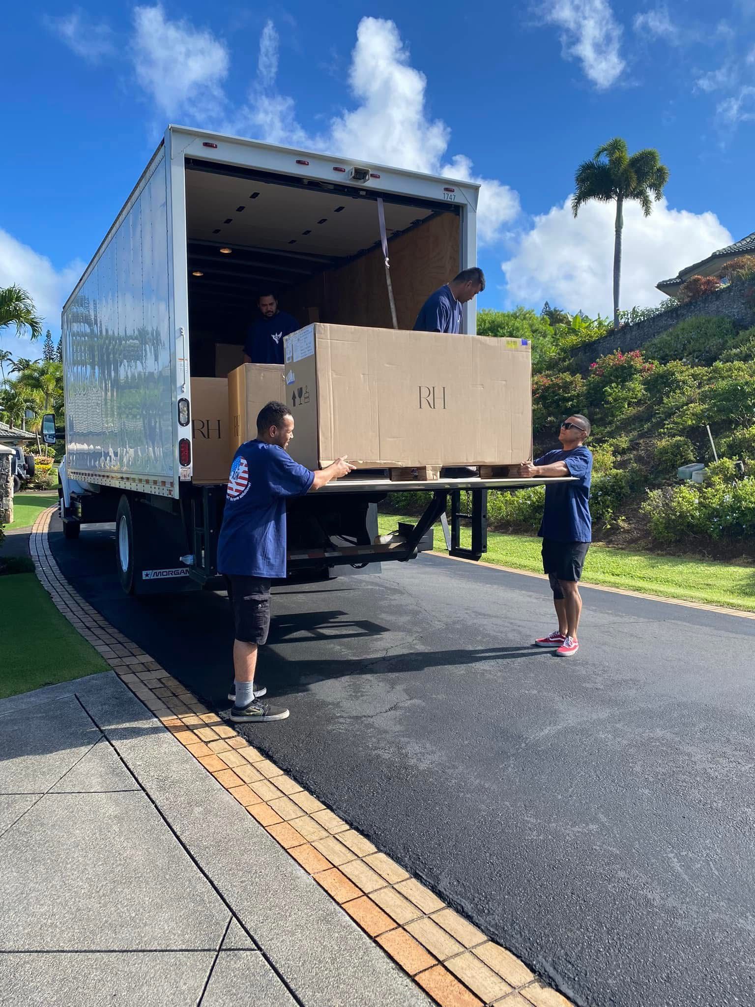 Movers loading a large cardboard box onto a truck from a driveway. They wear matching blue shirts and black shorts on a sunny day.