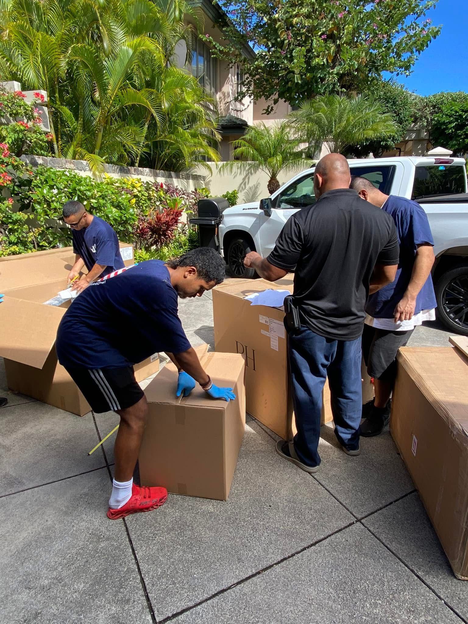 Men in blue shirts and a black shirt are unpacking boxes outdoors next to a white truck and tropical plants.