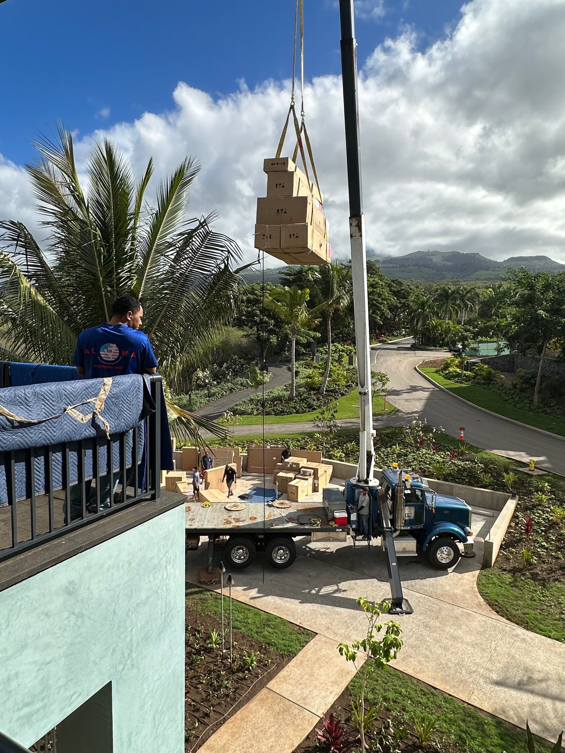 A crane lifts boxes onto a balcony. Workers on a truck and balcony observe the operation. Lush greenery and mountains are in the background.