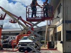 Two workers in a scissor lift near a building. A white pickup truck and other equipment are nearby.