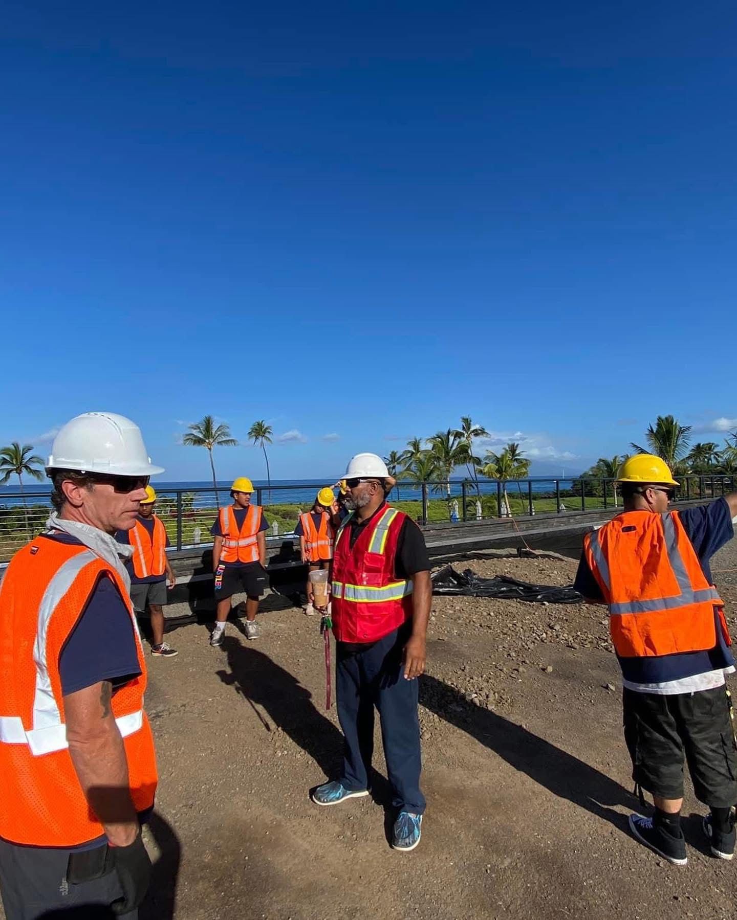 Construction workers in hard hats and safety vests standing outdoors, talking. Blue sky and ocean in the background.