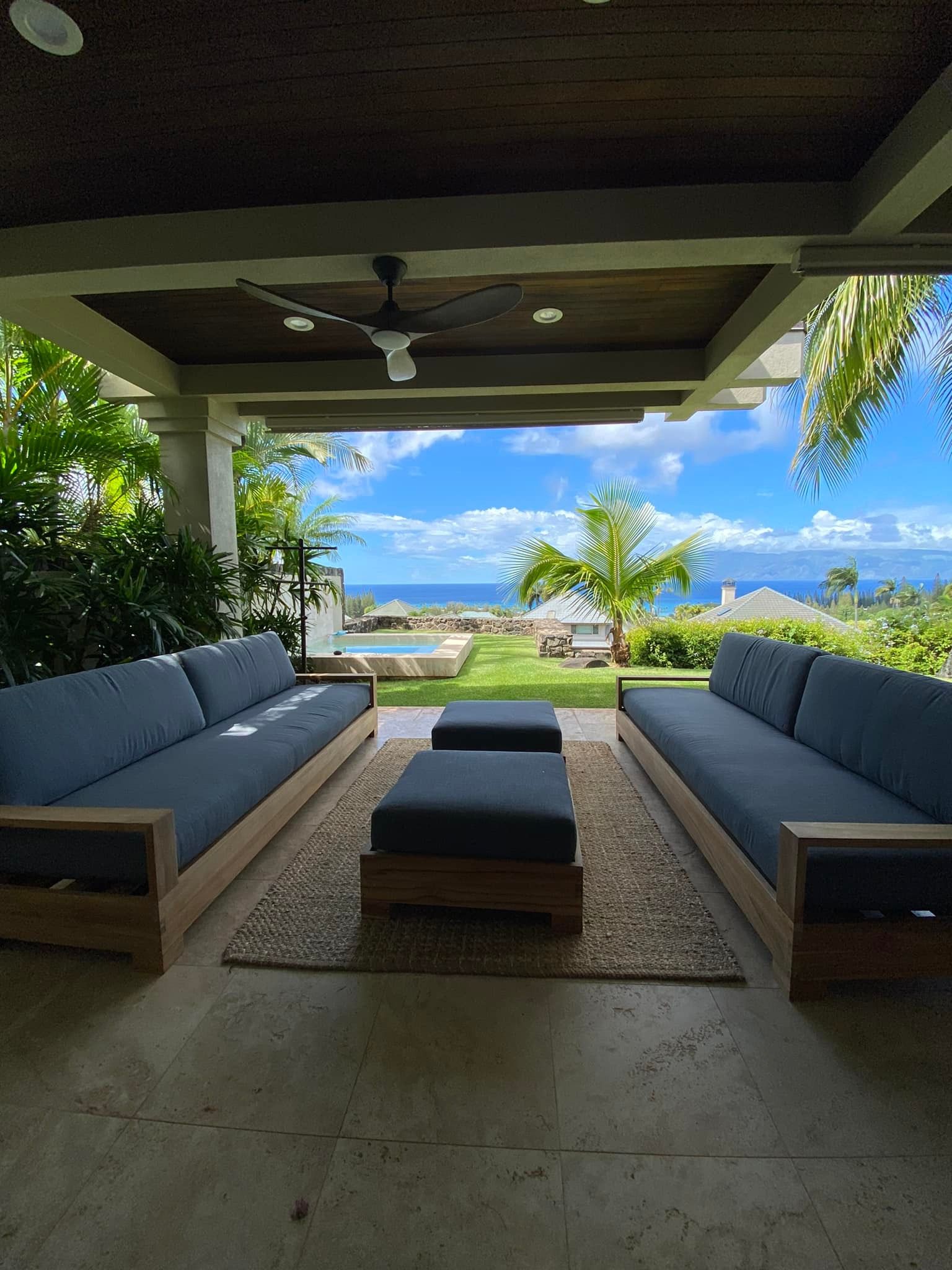 Outdoor patio with two blue-cushioned sofas and ottomans, overlooking a lawn, pool, and ocean under a blue sky.