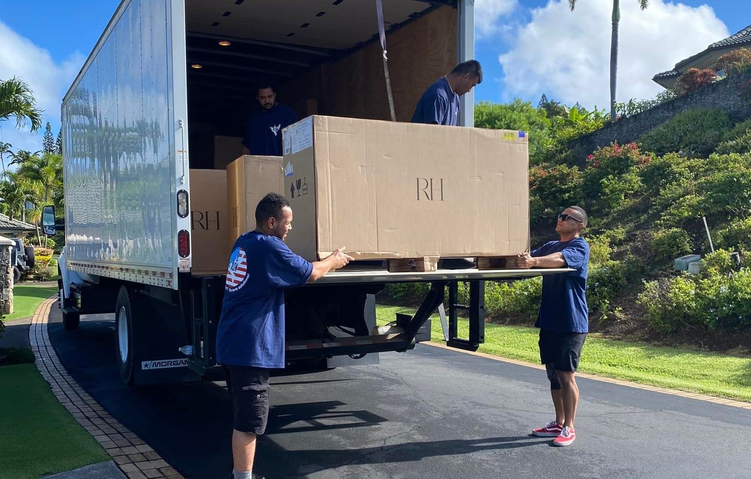 Movers loading a large box on a pallet into a truck on a driveway. Three movers are lifting the box, and one is in the truck.