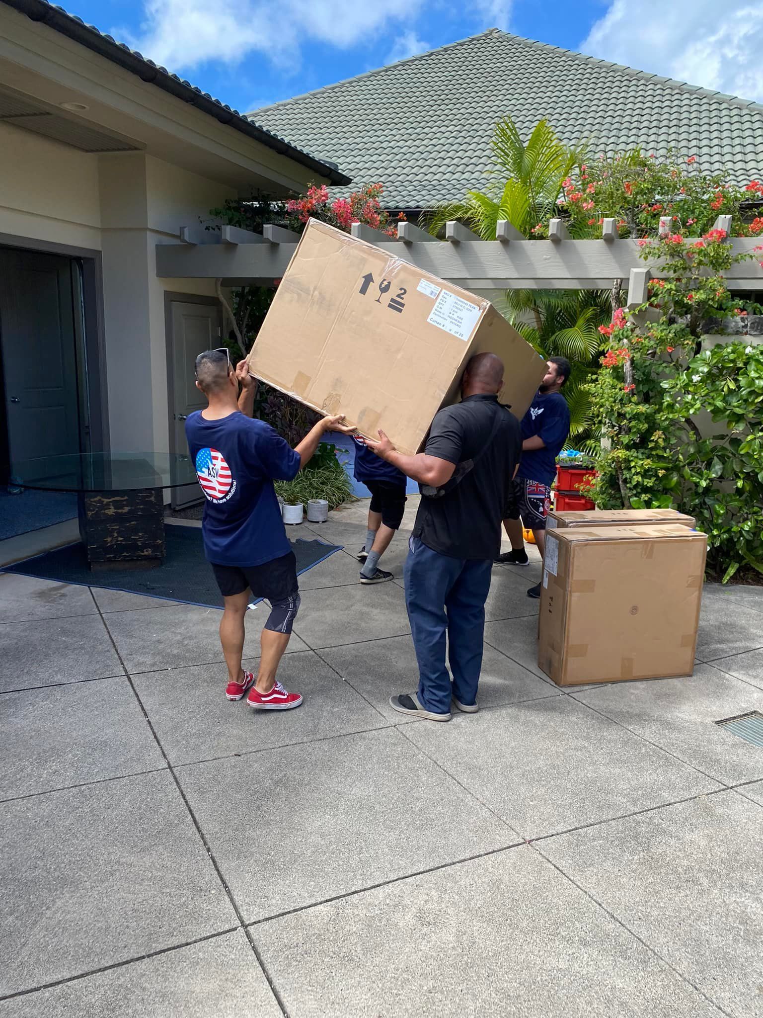 Several people lifting a large cardboard box outdoors on a patio. Others stand nearby, and there's foliage and a building in the background.