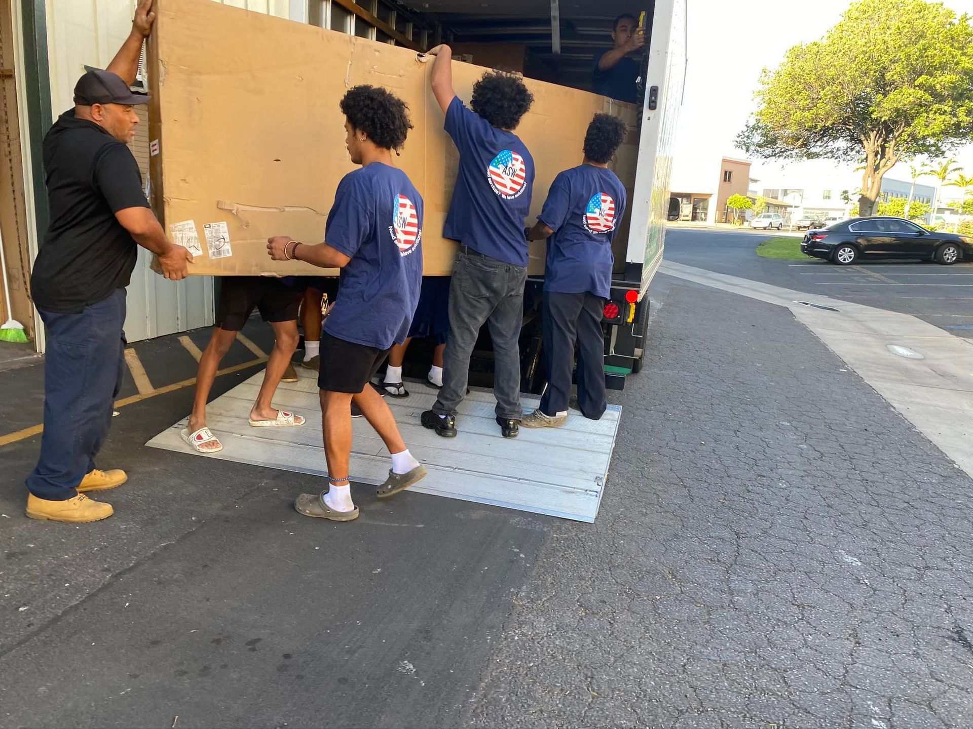 People loading a large cardboard item into a truck. Men and boys are outside in a parking lot, and one man is wearing a blue shirt with a flag on it.