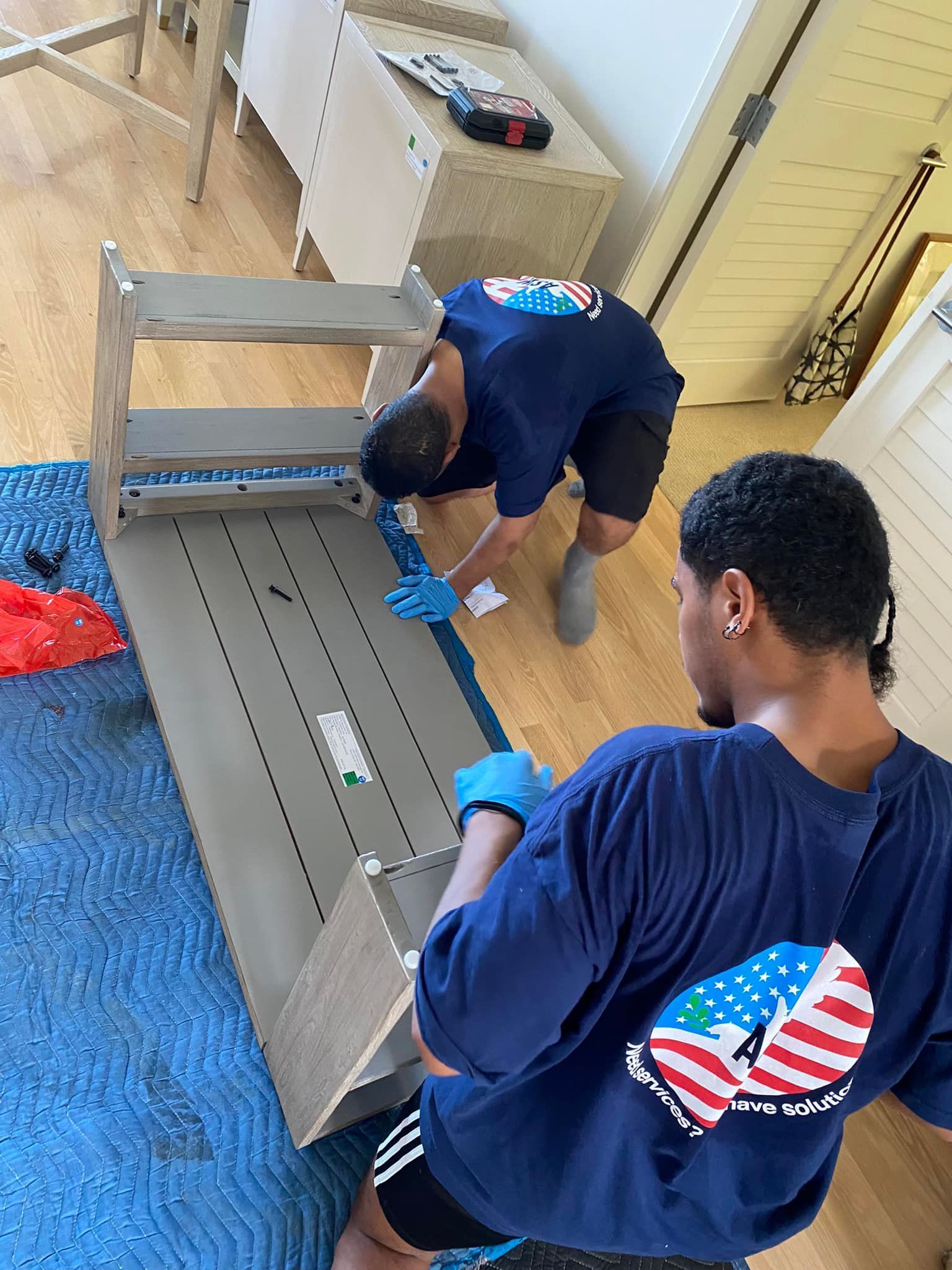 Two men in blue shirts with an American flag design assemble furniture on a blue tarp. They are wearing gloves and appear to be inside a house.