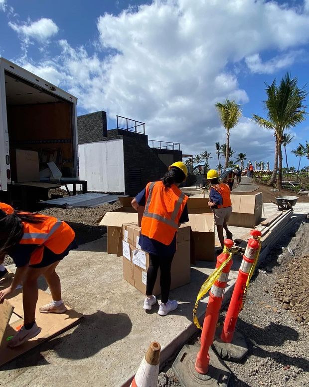 People in orange vests and hard hats unload boxes from a truck outdoors. Construction cones and palm trees are visible in the background.
