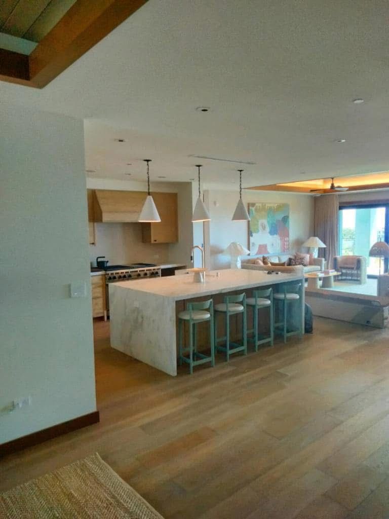 Spacious kitchen with a large marble island and teal bar stools. The room has hardwood floors and opens into a living area with a view.