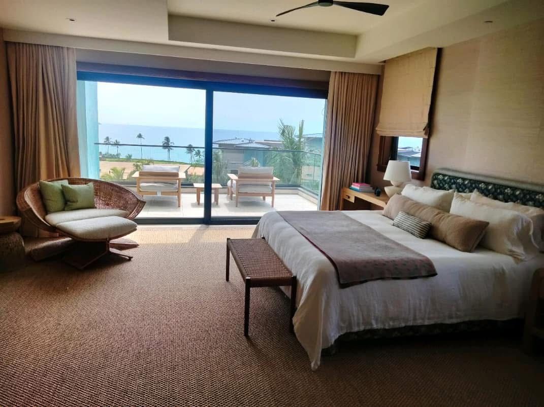Luxurious bedroom with a large bed, a lounge chair, and a balcony overlooking the ocean. The room has a neutral color palette and a rug.