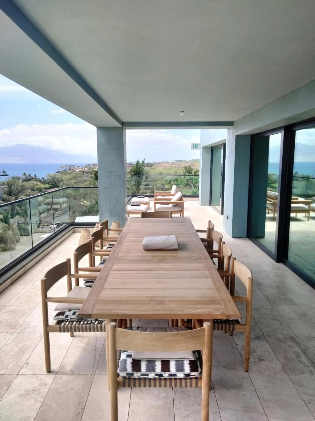 Outdoor dining table and chairs on a patio overlooking a coastal landscape, light-colored wood and beige stone, glass railing.