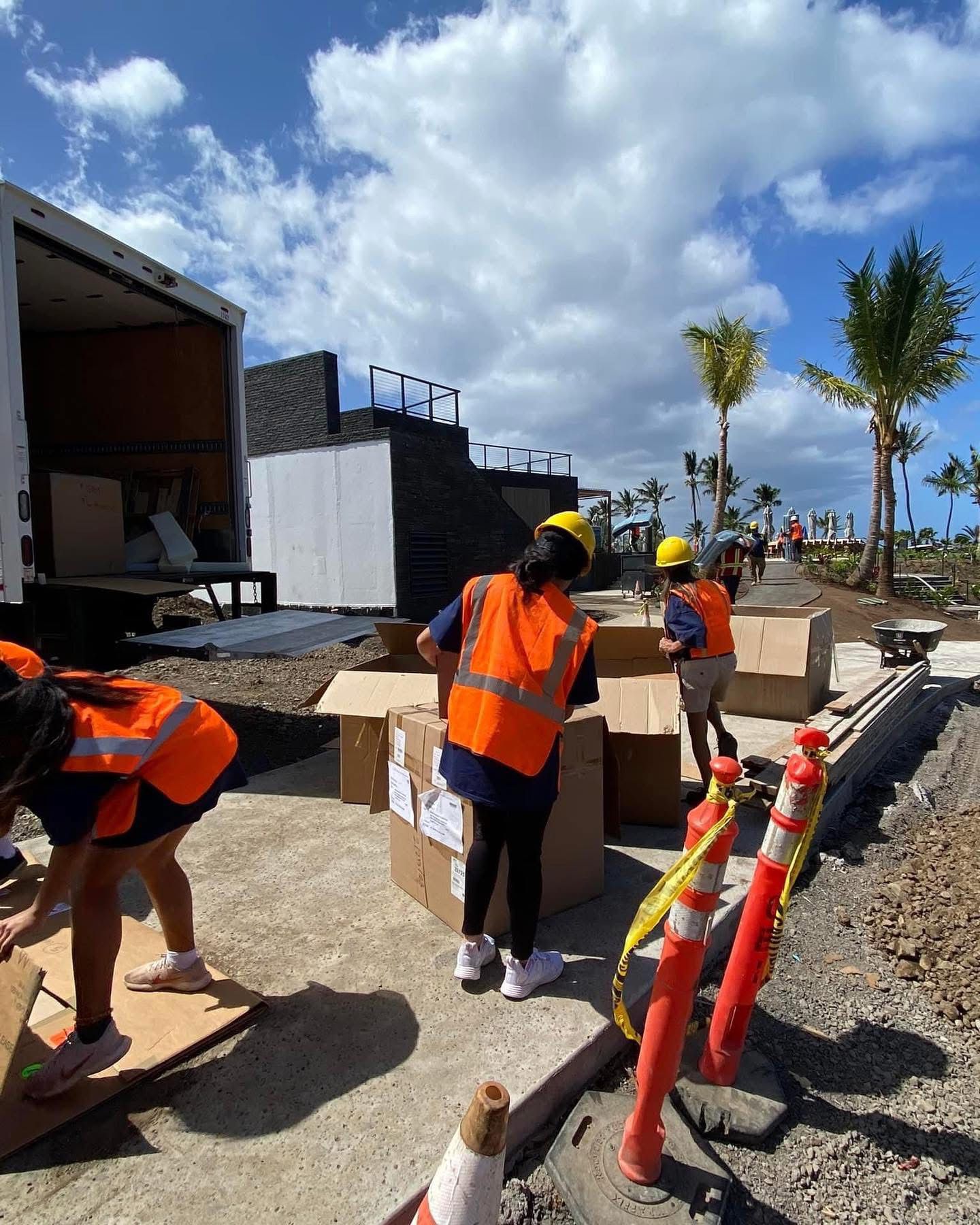 People in orange vests and hard hats unpack boxes at an outdoor construction site. A white truck and palms are in the background.
