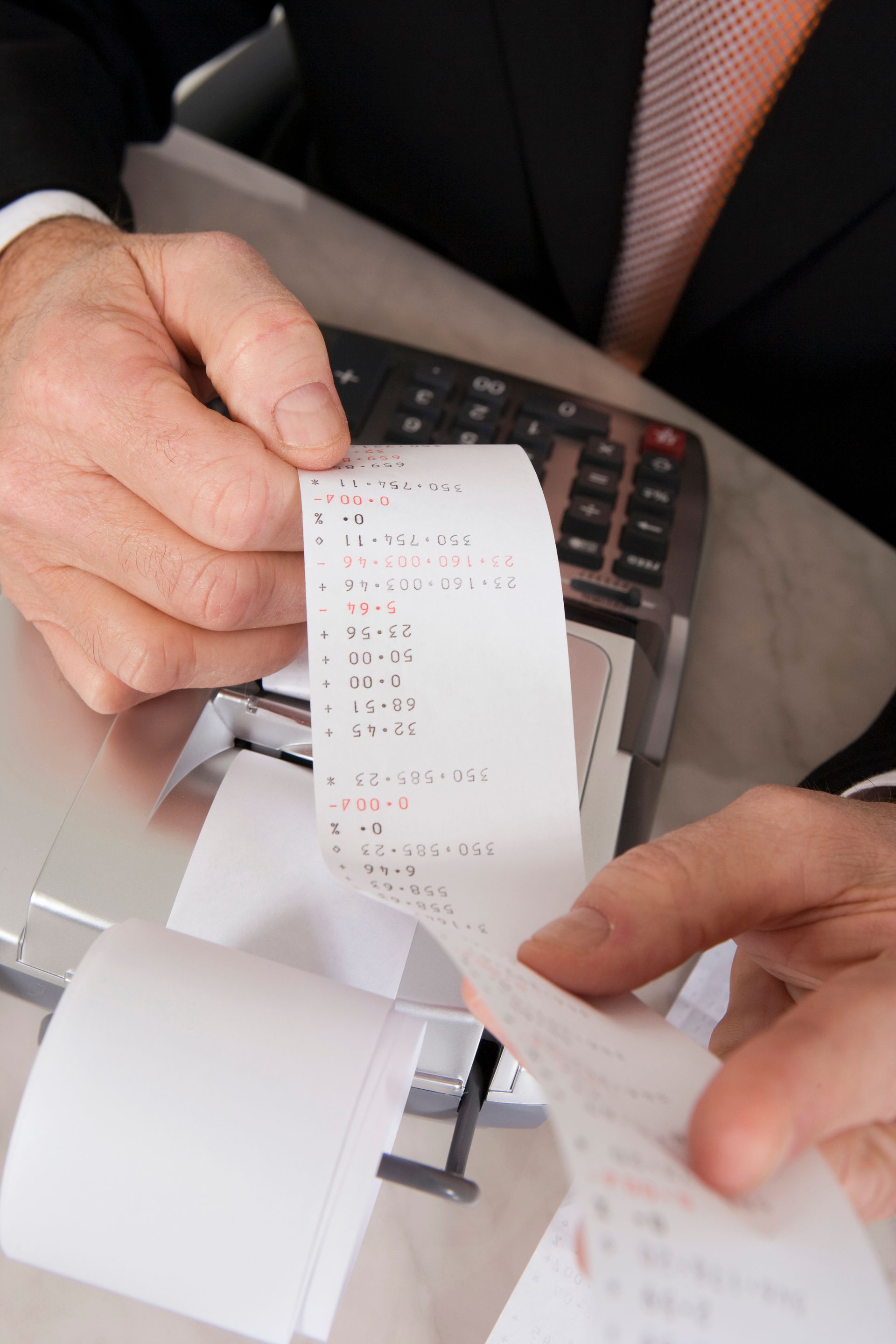 Hands holding and examining a long printed receipt from an adding machine, over a calculator.