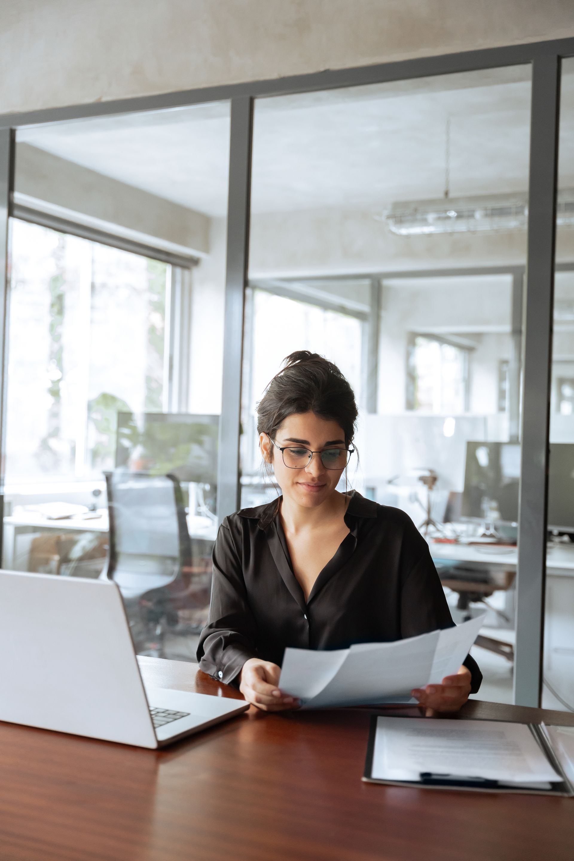 Woman in glasses reviews documents at a desk in a modern office.