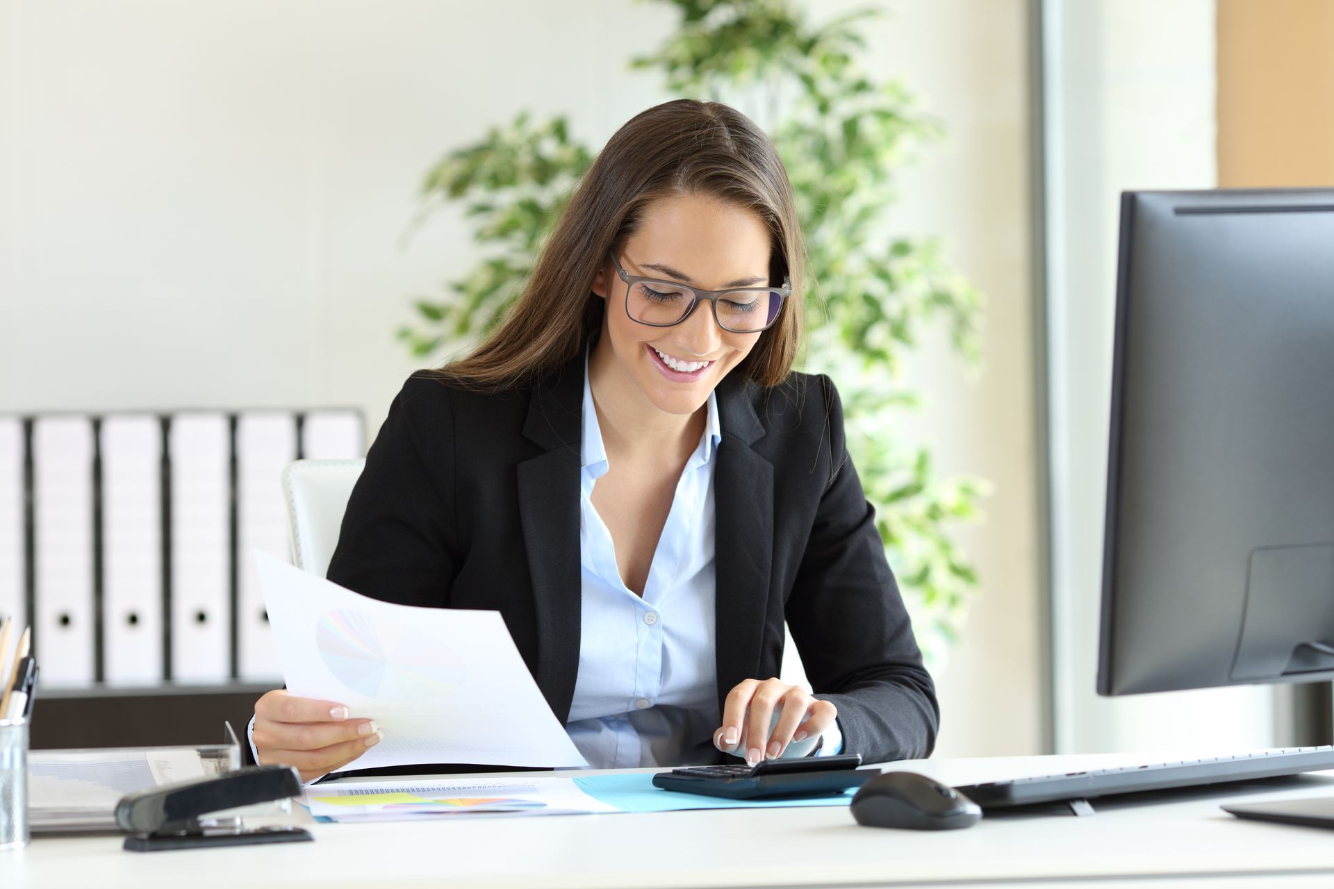 Woman in a suit smiling while working at a desk, looking at papers and using a calculator.