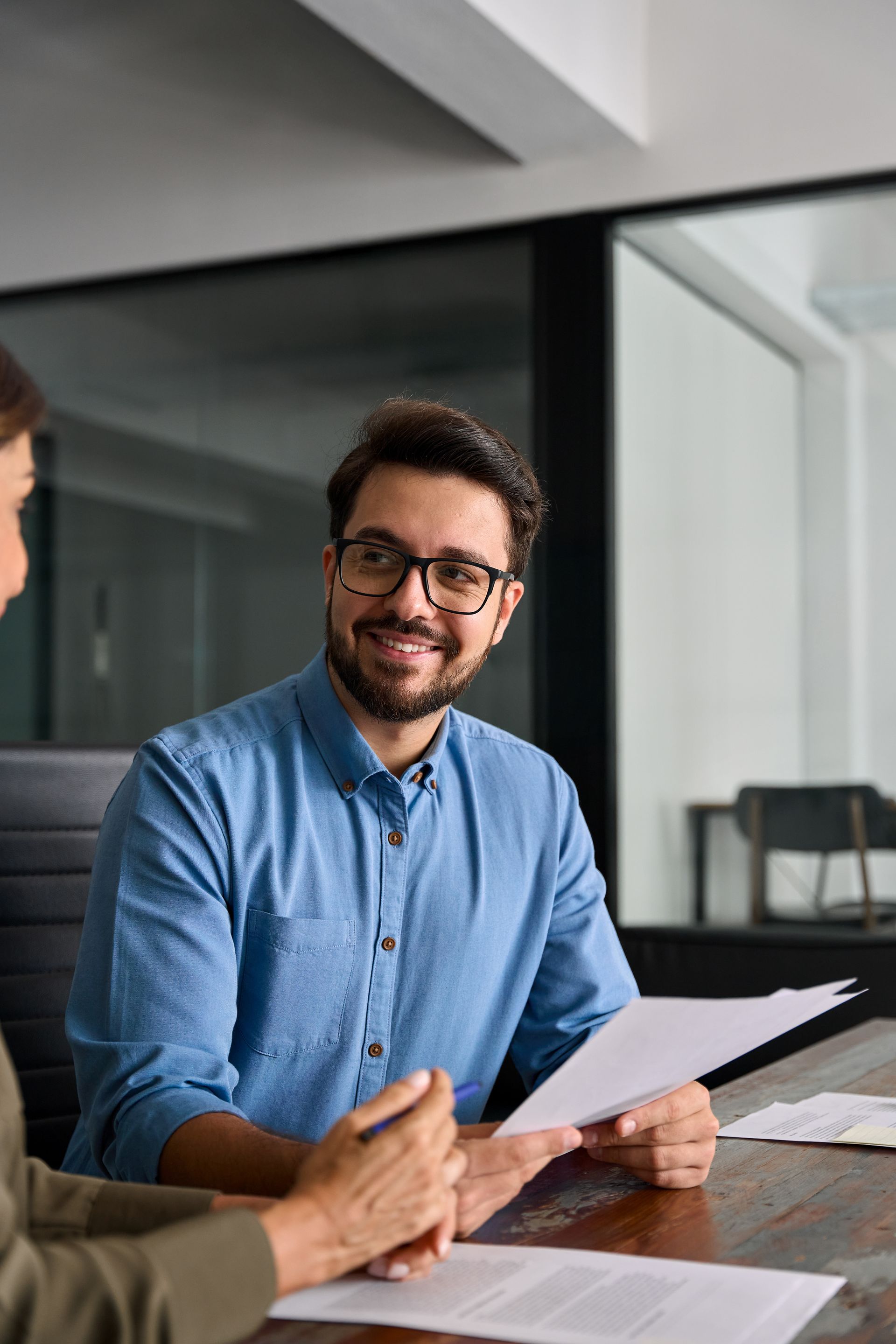 Man in blue shirt, glasses smiles while reviewing documents with another person at a table.
