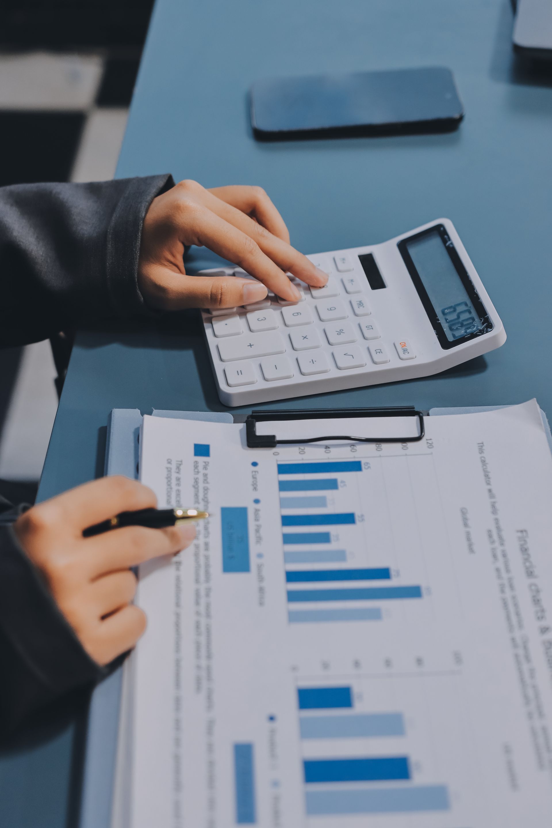 Hands using calculator and pen, analyzing financial charts on a desk.