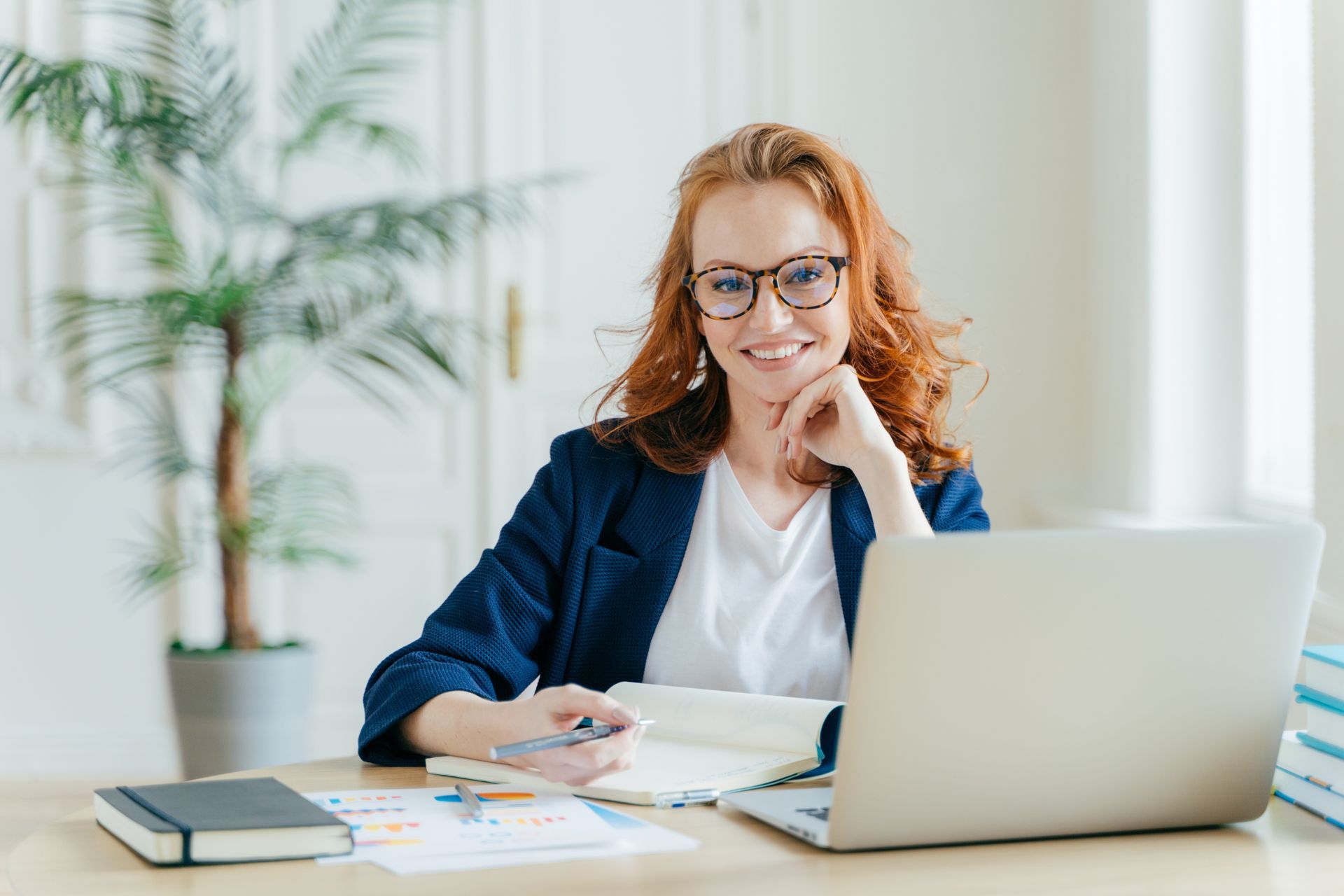 Woman with red hair and glasses smiles while working at a desk with a laptop and notebook.