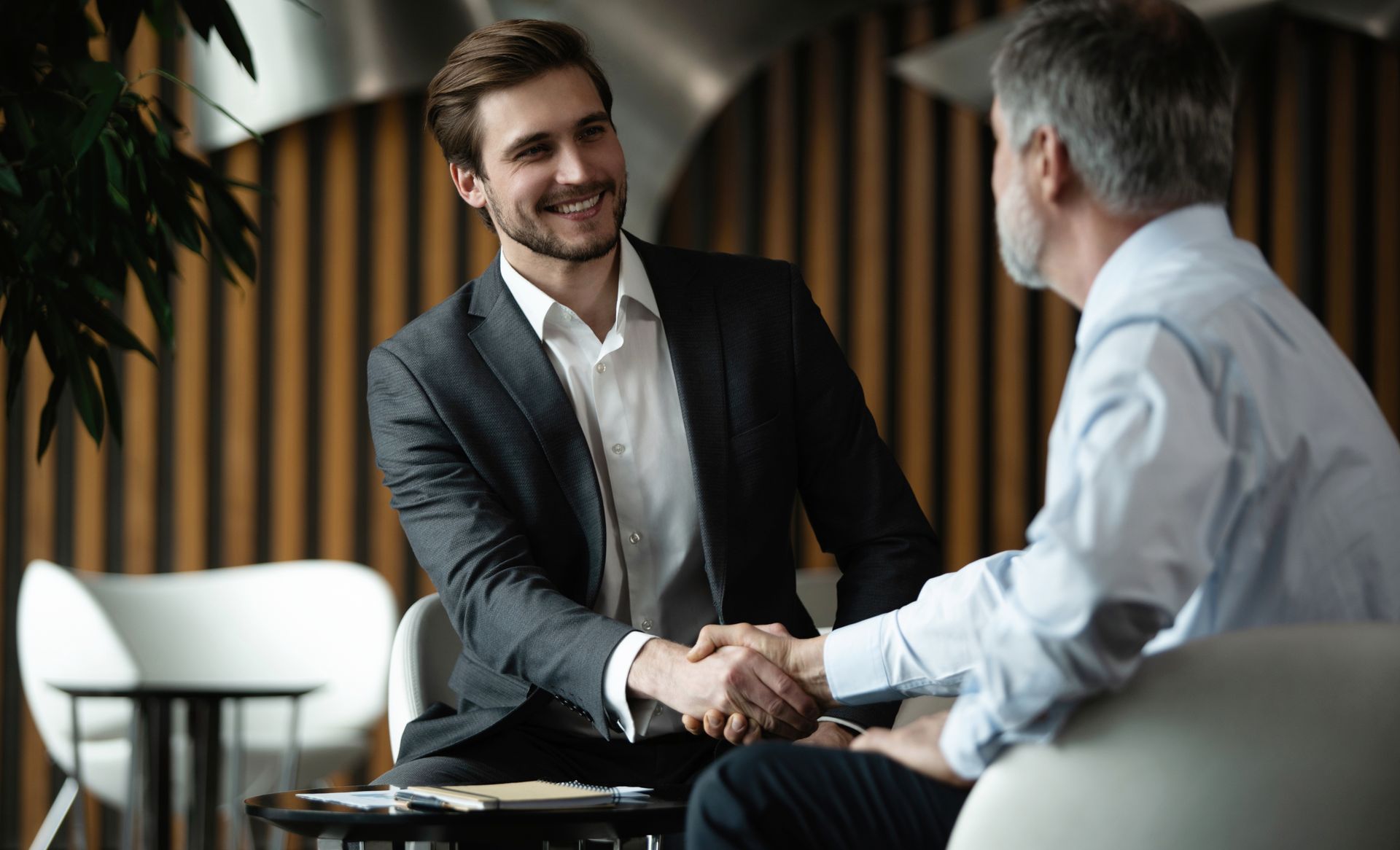 Two men in suits shaking hands, smiling, in an office setting.