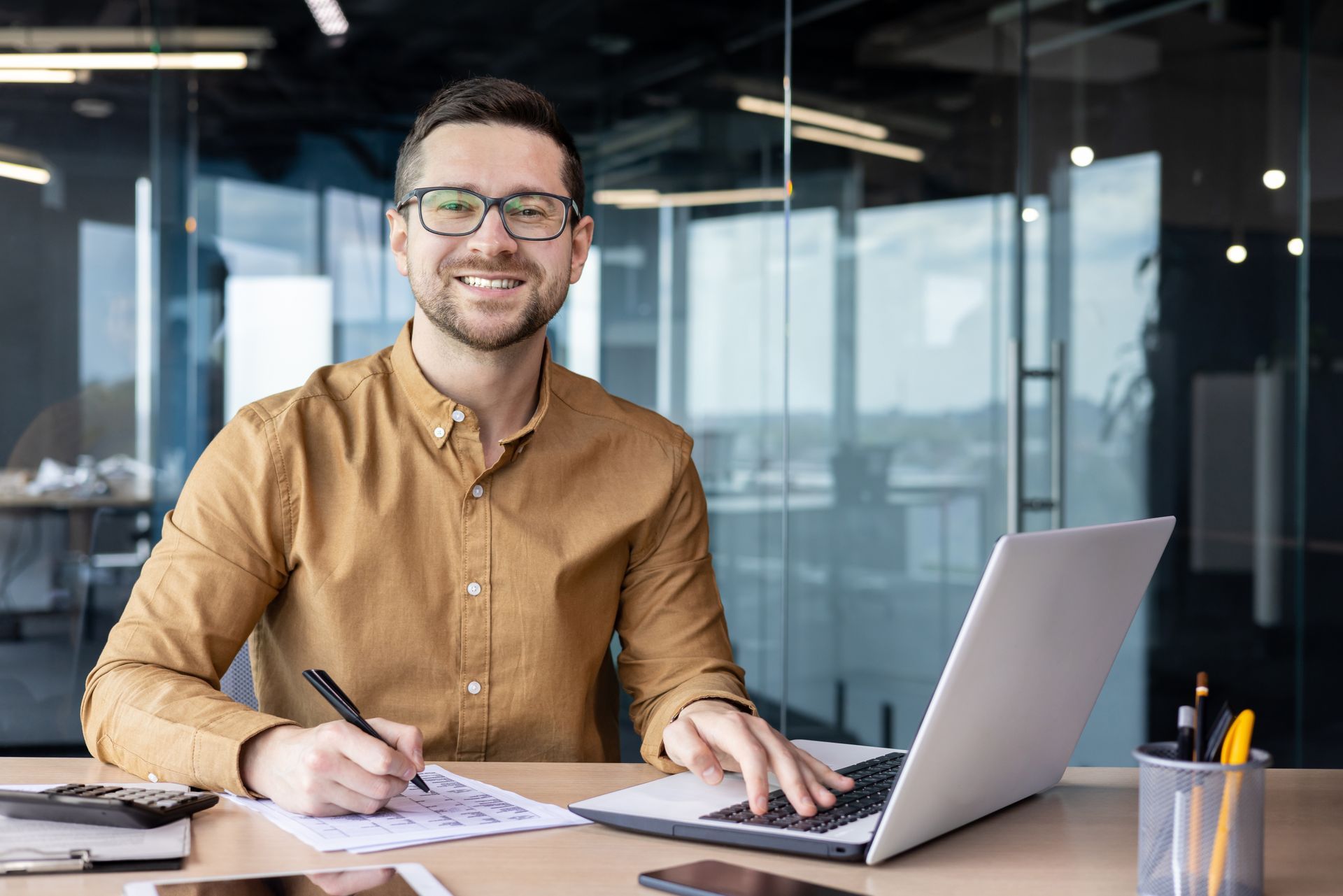 Man in glasses smiles at the camera, working on a laptop and writing in an office.