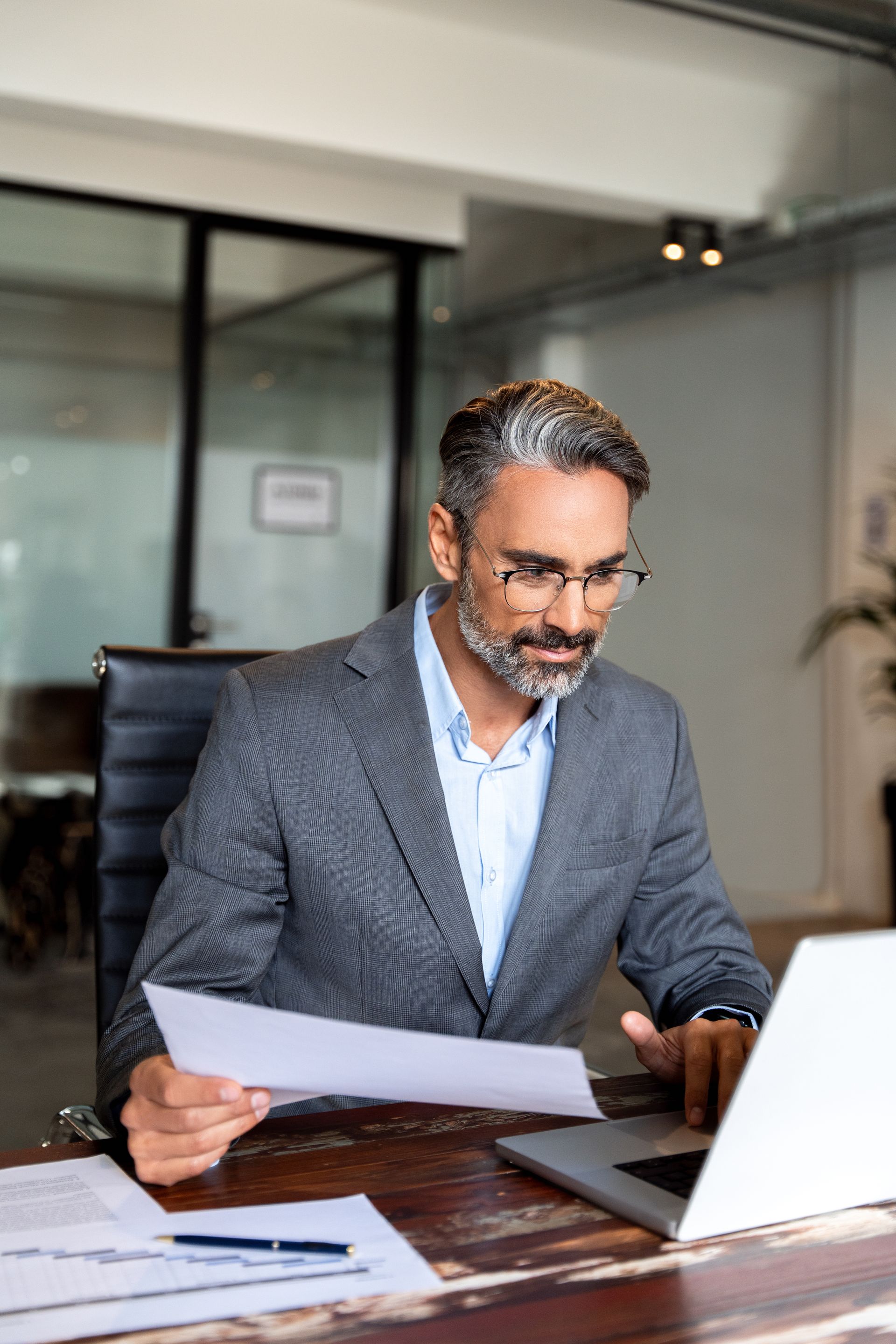 Man in a gray blazer working on a laptop, holding papers in an office setting.
